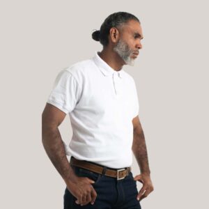 Un modelo masculino mayor con barba gris y cabello peinado hacia atrás, visto de perfil, usando una chemise polo blanca de manga corta sobre un fondo de estudio gris claro.